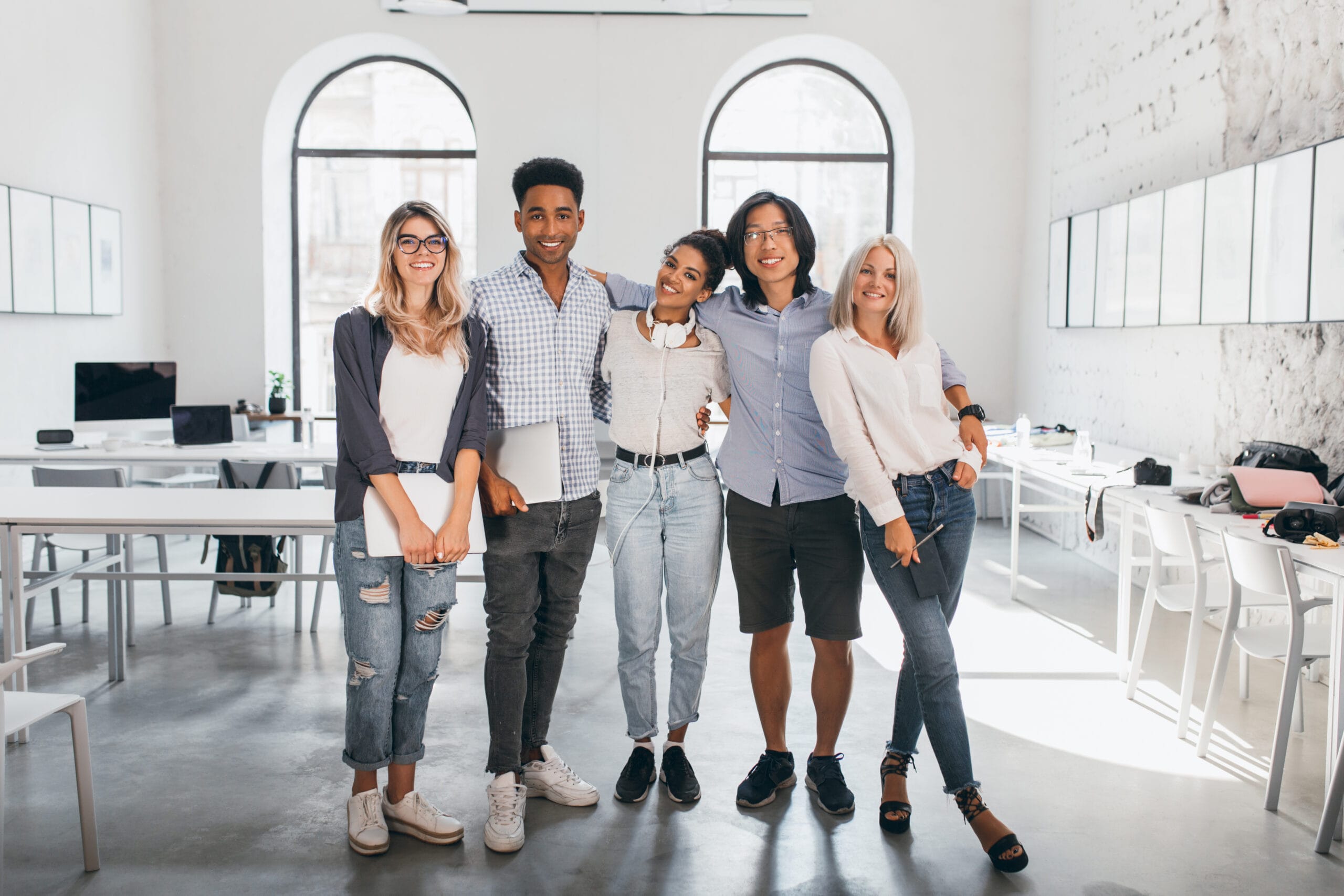 Full-length portrait of shy blonde girl in white sneakers holding laptop after seminar and stands beside african friend. Excited international students posing together after lecture in spacious hall..